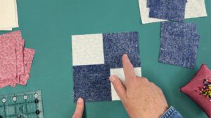 Hands pinning a four-patch quilt block with navy and cream fabric on a cutting mat